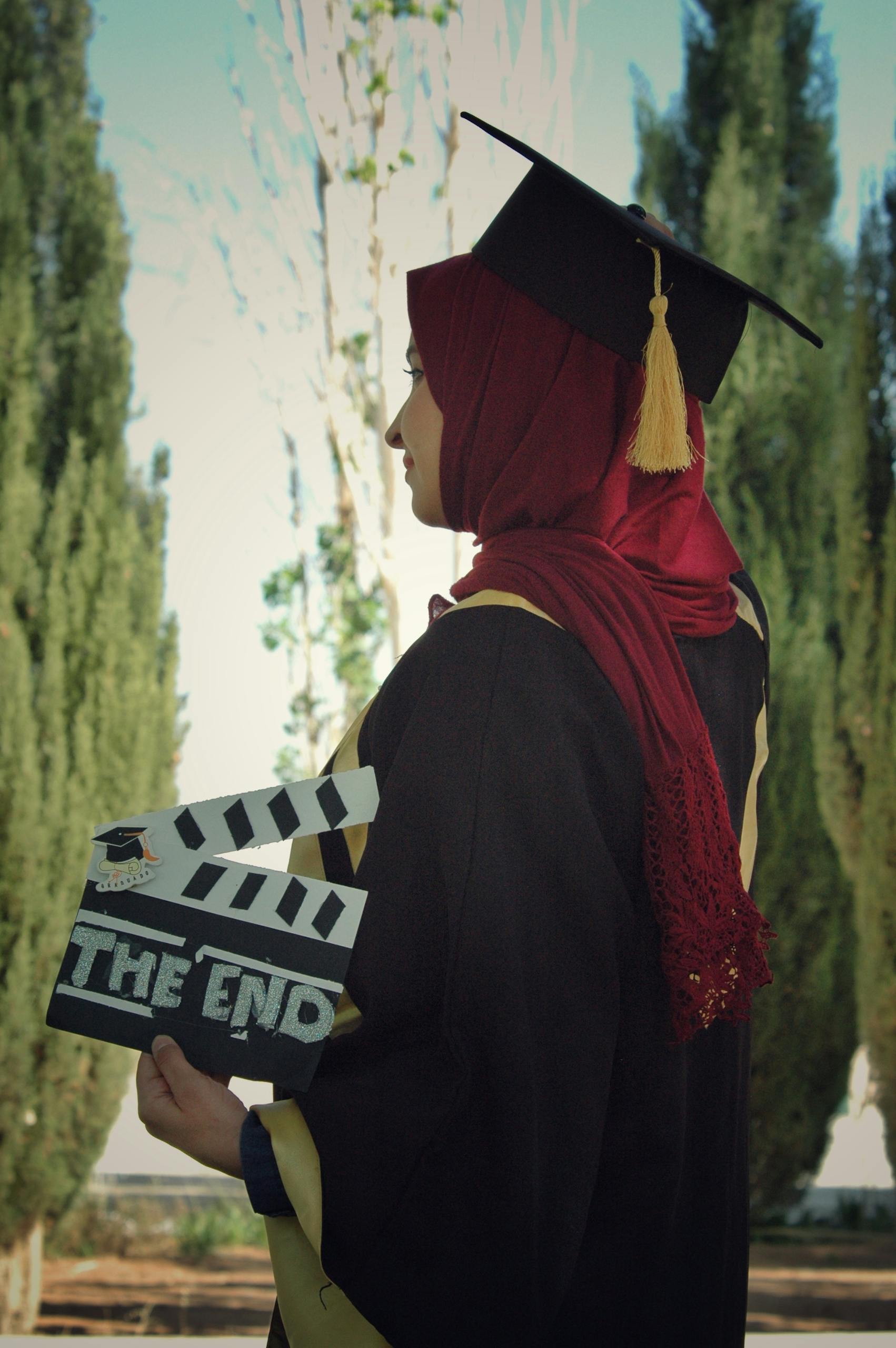 Side view of Muslim graduate in hijab holding clapperboard outdoors.
